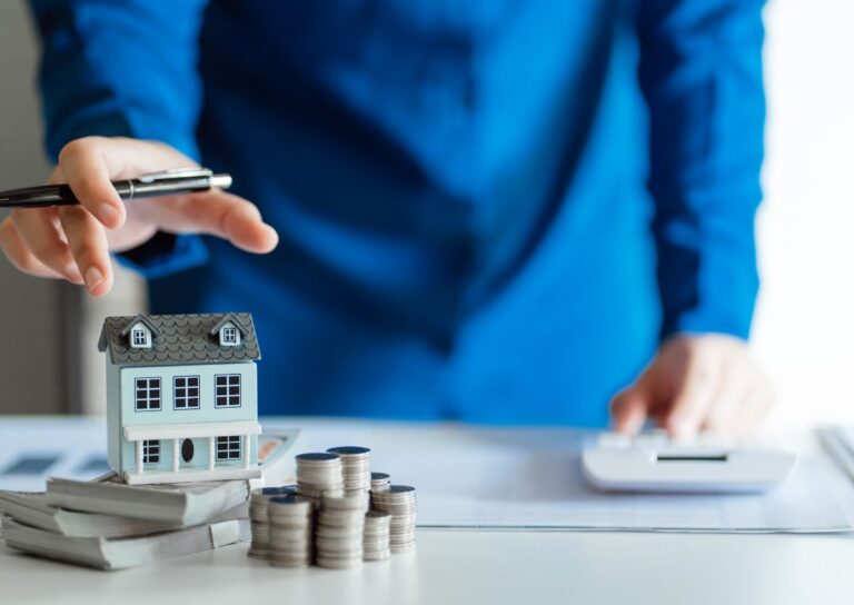 Person in blue shirt using calculator beside miniature house and stacked coins