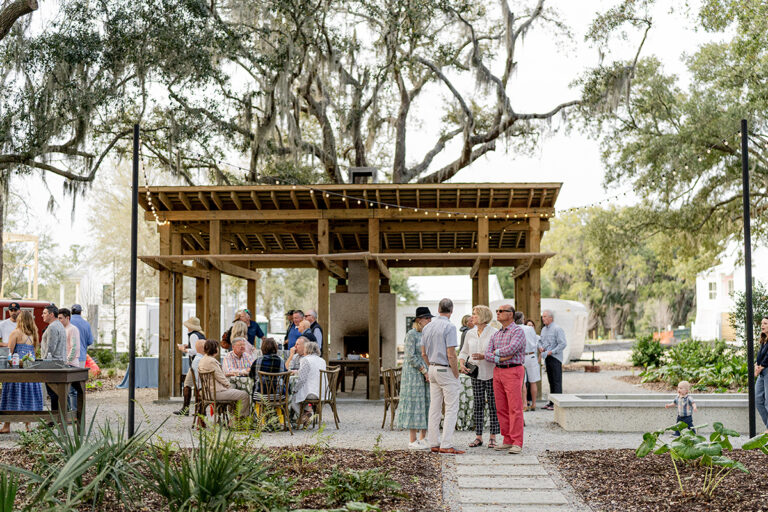 People seated under a rustic wood-beamed shelter in a garden setting.