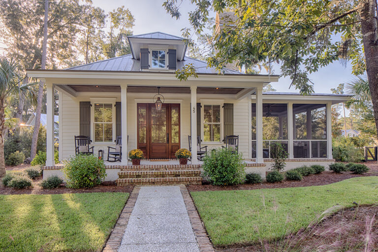 A charming Southern-style cottage with a wide front porch, rocking chairs, and symmetrical landscaping leading up to wooden double front doors.