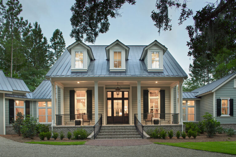 Front exterior view of a light-colored home with a metal roof, dormer windows, a covered front porch with columns and railing, steps leading up, and surrounding landscaping.