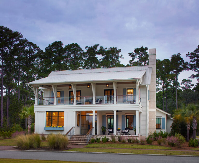 A white two-story residence with metal roofing, upper and lower porches, blue shutters, and a tall exterior chimney, framed by pine trees and manicured landscaping.