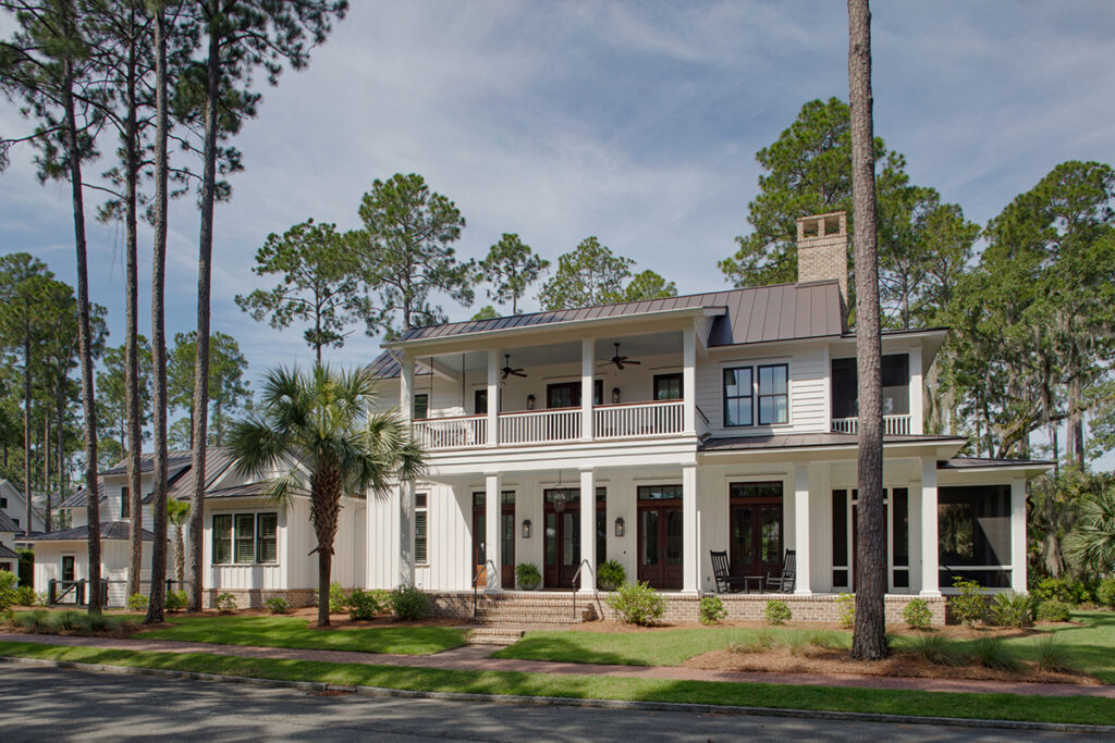 Front view of a white Southern-style home with a screened porch, standing seam metal roof, brick chimney, and lush green lawn surrounded by tall pine trees.