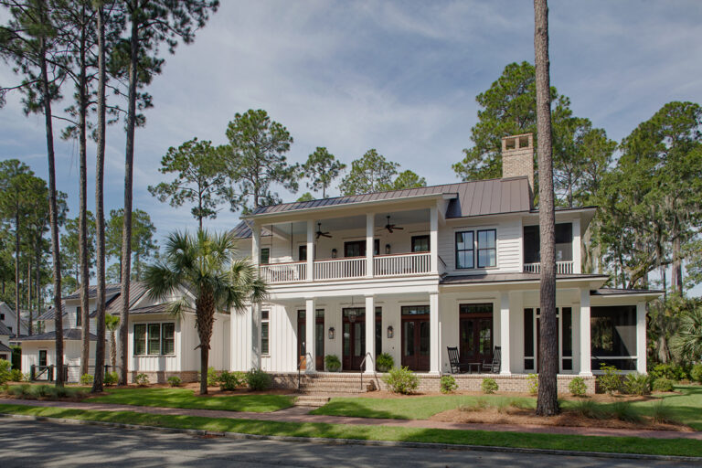 Front view of a white Southern-style home with a screened porch, standing seam metal roof, brick chimney, and lush green lawn surrounded by tall pine trees.