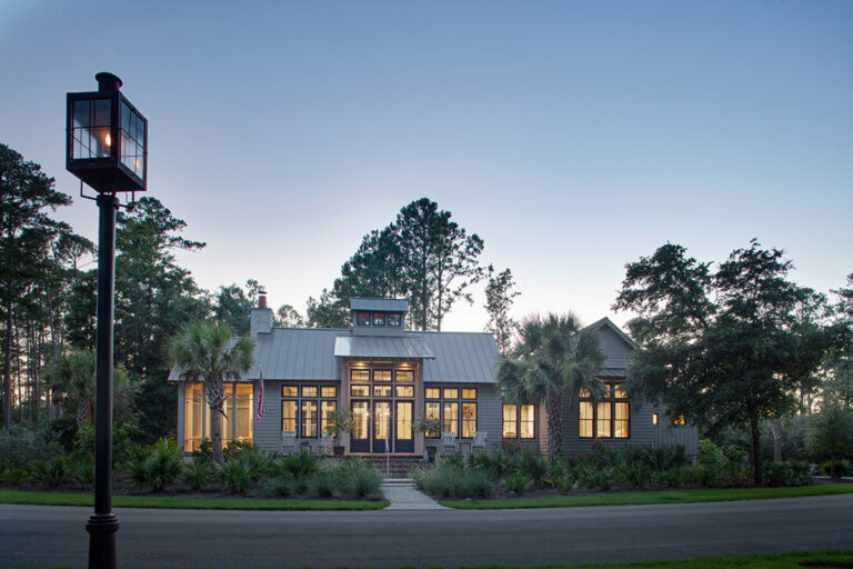 Exterior view of a modern grey home with a metal roof, large windows, and a central glass section, illuminated from within at dusk, with a traditional gas lantern in the foreground.